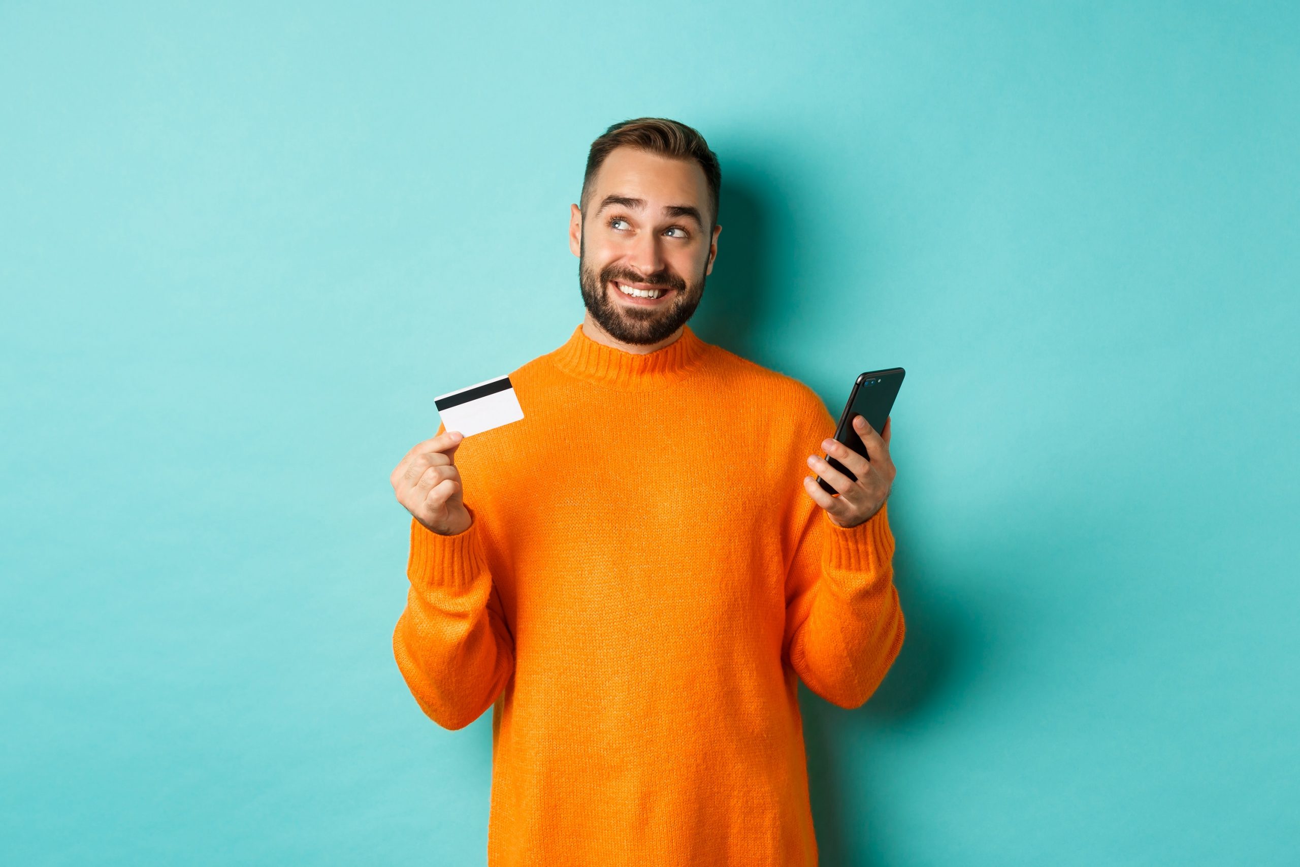 Online shopping. Handsome man thinking, holding smartphone with credit card, paying in internet store, standing over light blue background
