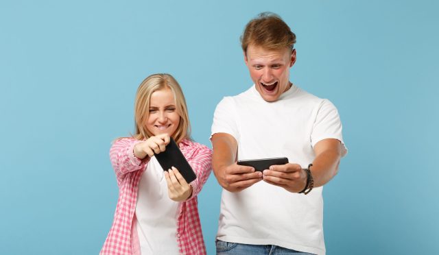 Young cheerful couple two friends guy girl in white pink blank t-shirts posing isolated on pastel blue background. People lifestyle concept. Mock up copy space. Playing video game with mobile phones.