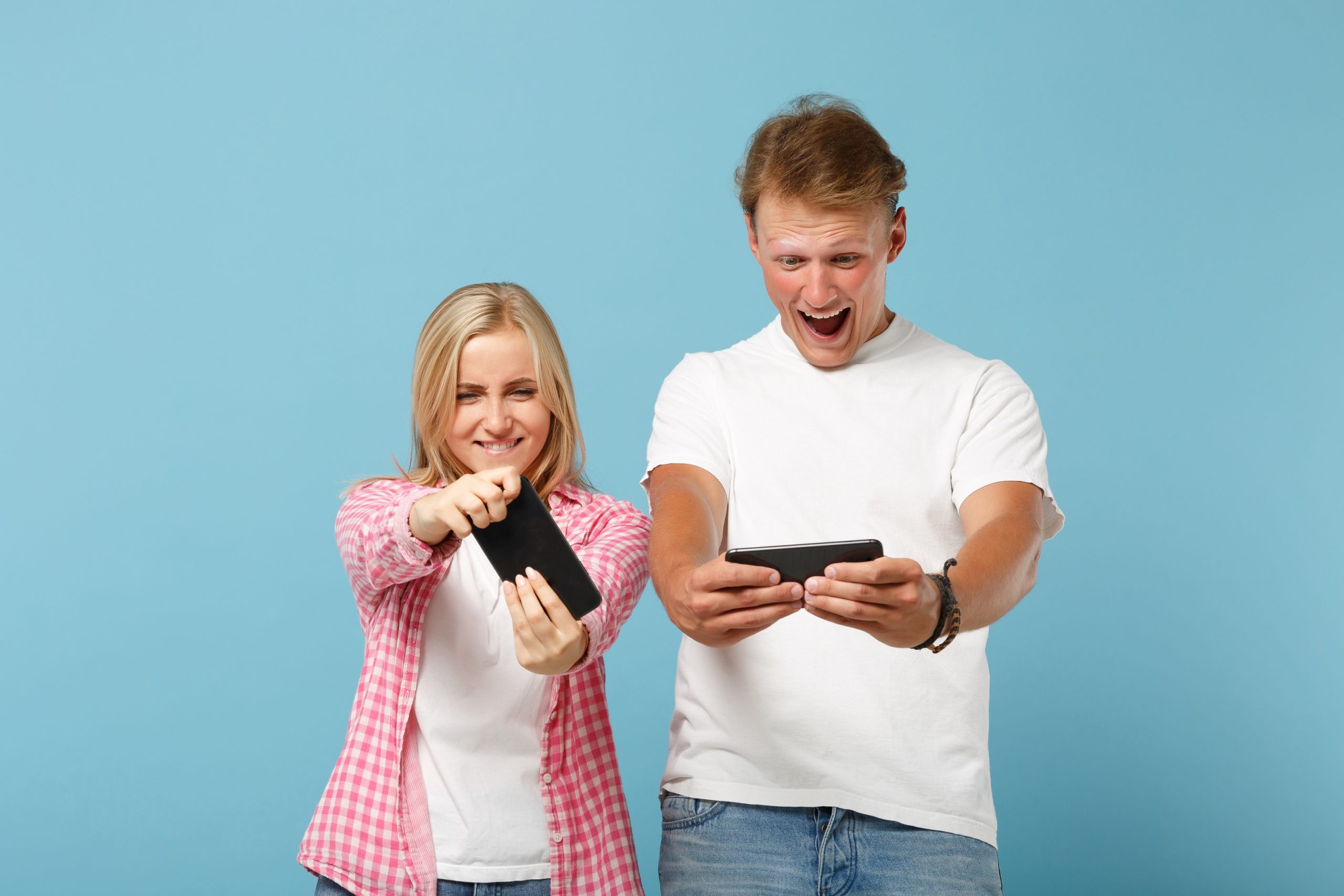 Young cheerful couple two friends guy girl in white pink blank t-shirts posing isolated on pastel blue background. People lifestyle concept. Mock up copy space. Playing video game with mobile phones.